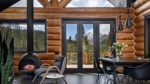 A modern log cabin interior with a fireplace, dining area, and large windows offering mountain views.