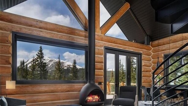 A modern log cabin interior with a fireplace, large windows, and a staircase.