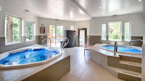 Un espace piscine intérieur moderne, doté de deux bains à remous, de grandes fenêtres et offrant une vue sur l’extérieur.