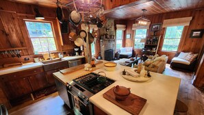 Interior rústico de una cabaña con una cocina que incluye estufa, horno y diversos utensilios de cocina. Hay una zona de comedor con mesa y sillas, y un rincón de estar acogedor con sofá y sillones.