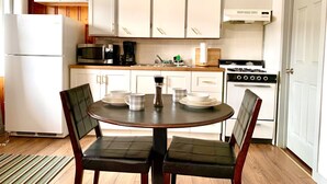 A kitchen with white cabinets, a stove, a microwave, and a refrigerator. There is a dining table with chairs and a coffee maker.