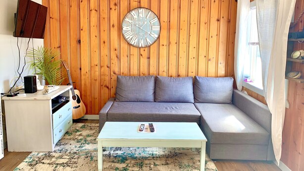 A living room with a wooden paneled wall, a round clock, a grey sofa, a white coffee table, and a TV on a stand.