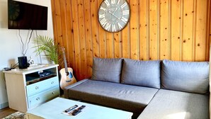 A living room with a wooden panel wall, a grey sofa, a white coffee table, a guitar, and a TV on the wall.