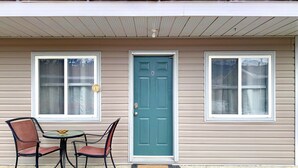 A single-story house with a front door, two windows, a small table with chairs, and a doormat.