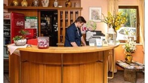 A reception desk with a man standing behind it. There is a Coca-Cola refrigerator, a vase with flowers, and a window with curtains. - Zum kleinen Reichstag (Moers)