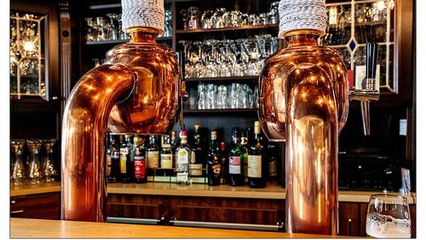 Two copper-colored bar taps with a textured cap, set against a backdrop of shelves with various bottles and glasses. - Zum kleinen Reichstag (Moers)