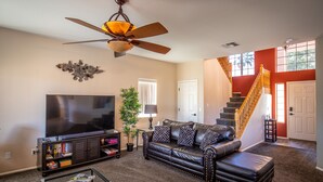 A living room with a leather sofa, a flat-screen TV, a bookshelf, a coffee table, and a ceiling fan. - Goodyear, AZ full home with heated pool and spa and desert oasis back yard!! (Goodyear)