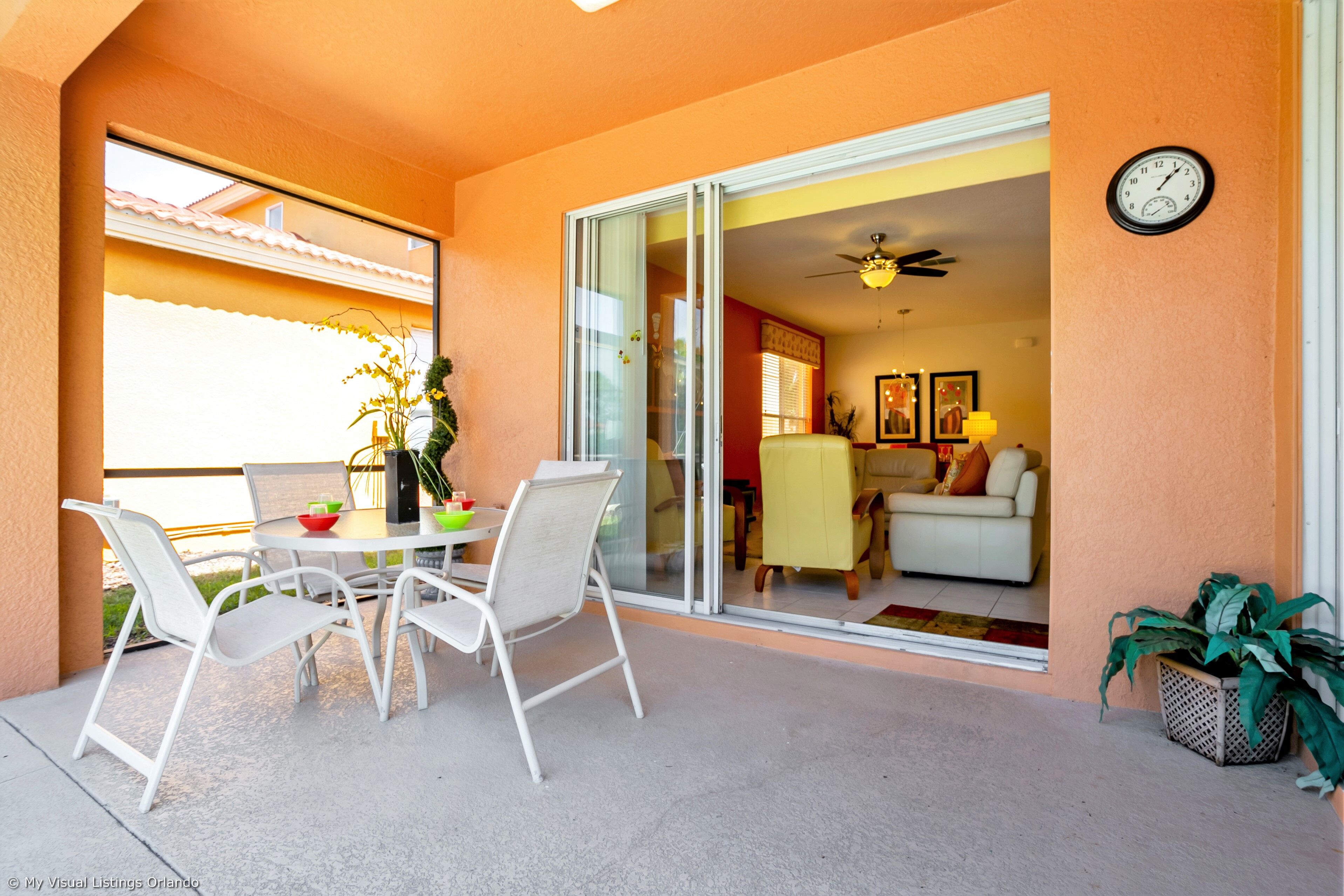 Um pátio com mesa e cadeiras, uma porta de vidro deslizante que dá para uma sala de estar e um relógio na parede.