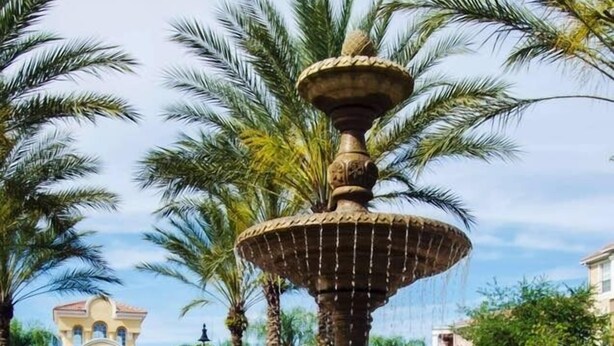 Une fontaine en pierre à plusieurs niveaux, dont l’eau jaillit en cascade, est entourée de palmiers et, au fond, on aperçoit un bâtiment avec un toit en tuiles.