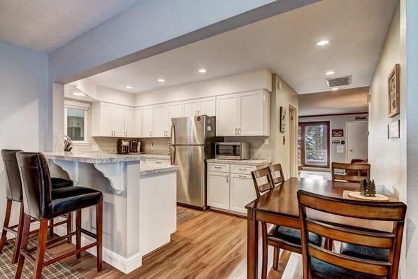 Una cocina con armarios blancos, un refrigerador de acero inoxidable, un microondas y un comedor con sillas de madera y una mesa.