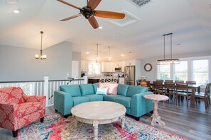 Un salon spacieux avec un canapé de couleur turquoise, une table basse ronde et un fauteuil à motifs rouges. On peut voir un ventilateur de plafond et des lampes suspendues.