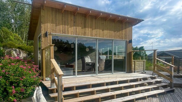 Une cabane en bois avec une porte d’entrée en verre, une terrasse en bois et un espace salon extérieur.