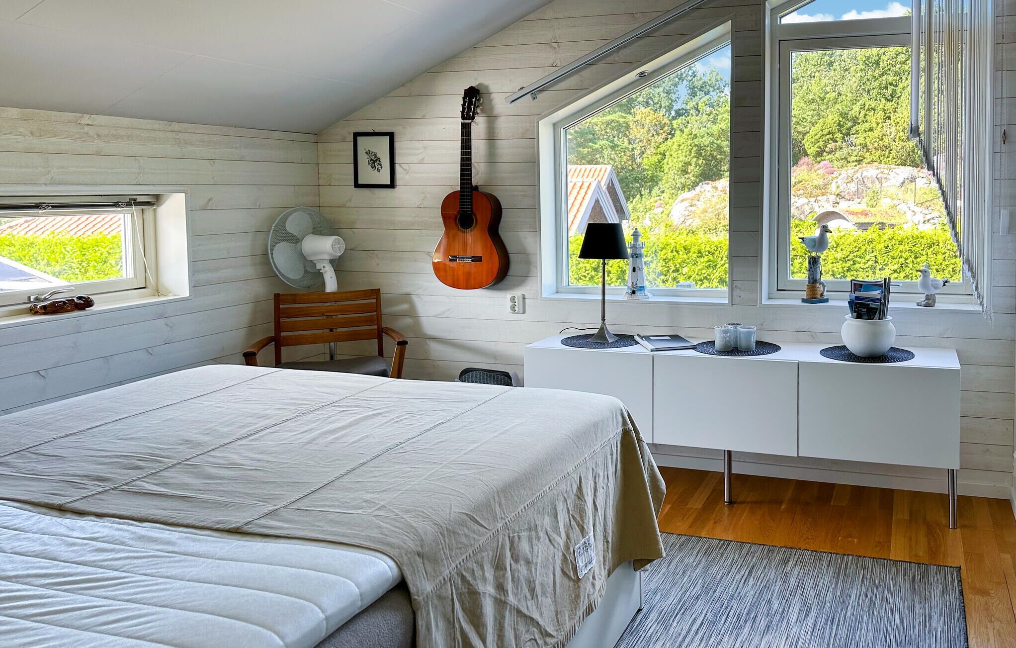 Une chambre à coucher avec un lit, une guitare, un bureau avec une lampe et une fenêtre donnant sur un paysage verdoyant.