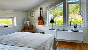 Une chambre à coucher avec un lit, une guitare, un bureau avec une lampe et une fenêtre donnant sur un paysage verdoyant.