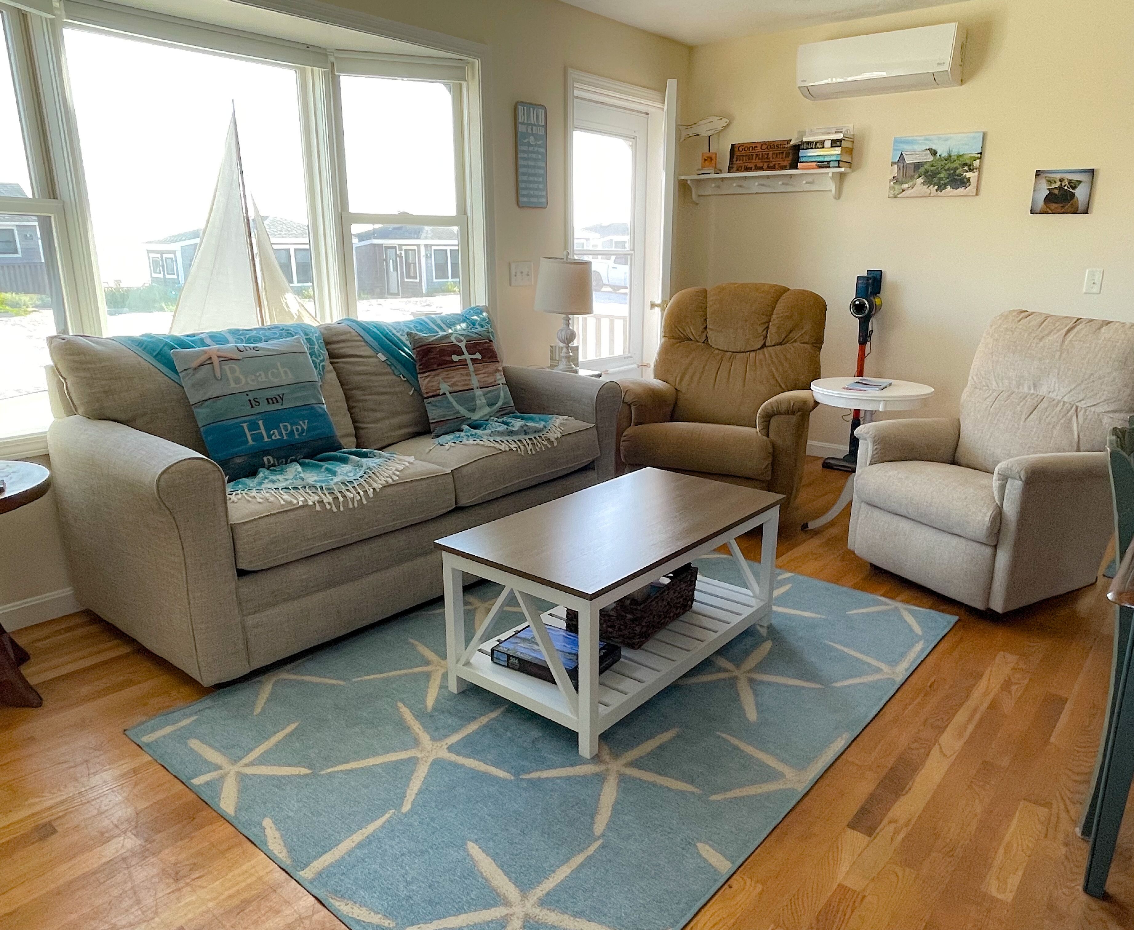 Een woonkamer met een beige bank, een salontafel en twee fauteuils. Er is een raam met uitzicht op huizen.