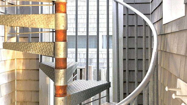 A spiral staircase with metal railings and a central pole, leading to a balcony with a view of a building with wooden siding. - Lovely Oceanview 2 Bed 2 Bath with sleeper sofa, outdoor shower & rooftop deck! (North Truro)