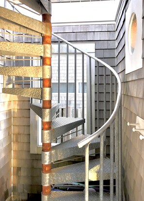 Un escalier en colimaçon avec des rampes en métal et un pilier central, menant à un balcon d’où l’on peut admirer un bâtiment au revêtement en bois.