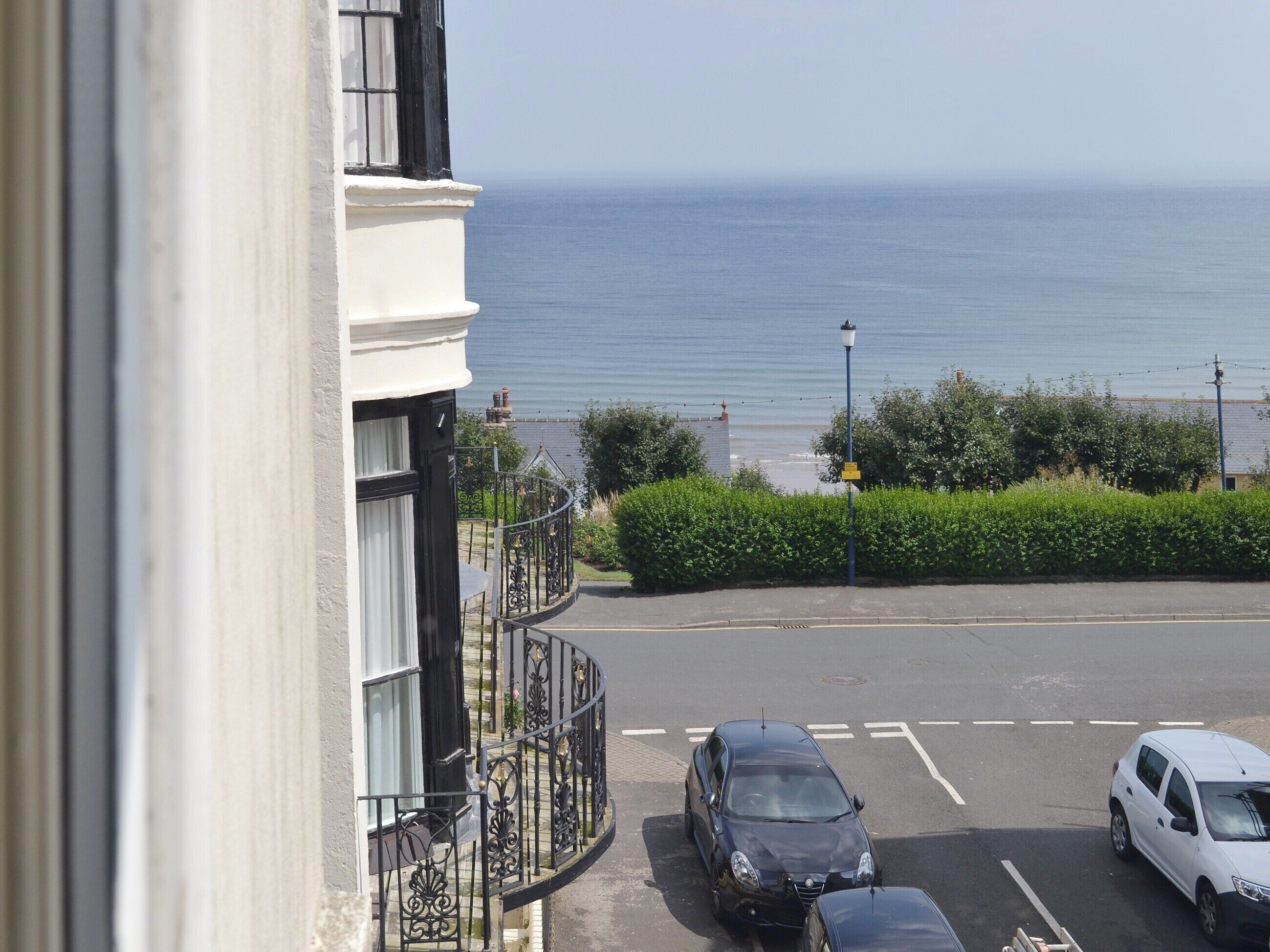 A seaside view from a balcony overlooking a parking area with cars, a hedge, and a distant shoreline.