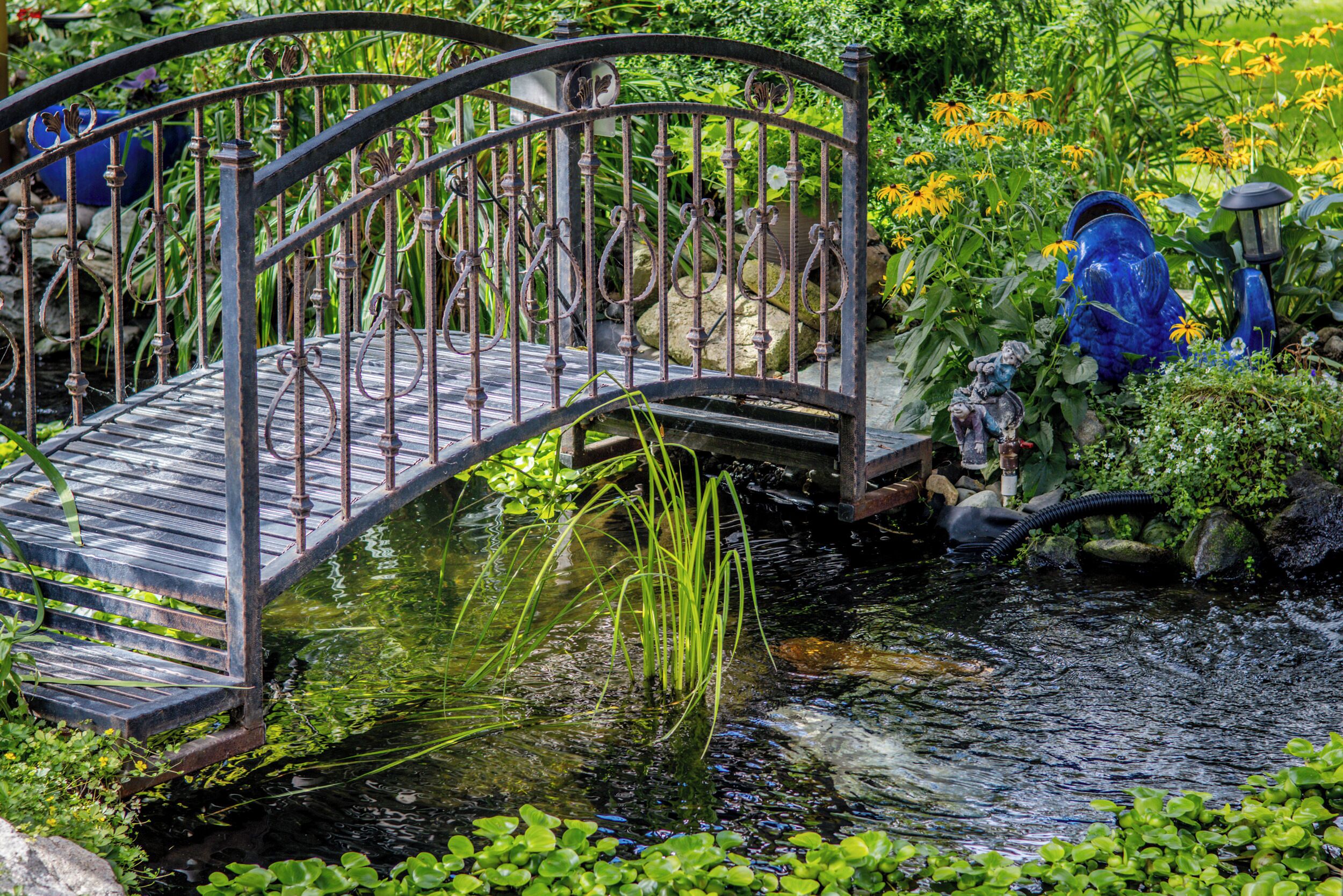 A metal bridge over a pond with water lilies and a statue.