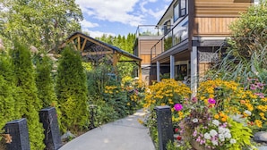 A modern house with a wooden deck, a concrete pathway, and a garden with various flowers. - Rayleigh Riverfront Retreat -BEAUTIFUL GARDEN OASIS (Kamloops)