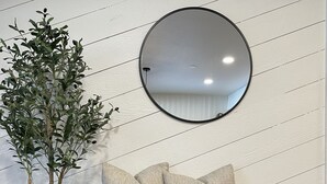 Eine Holzbank mit zwei Kissen, einer Topfpflanze und einem runden Spiegel an einer Wand mit diagonalen Linien.