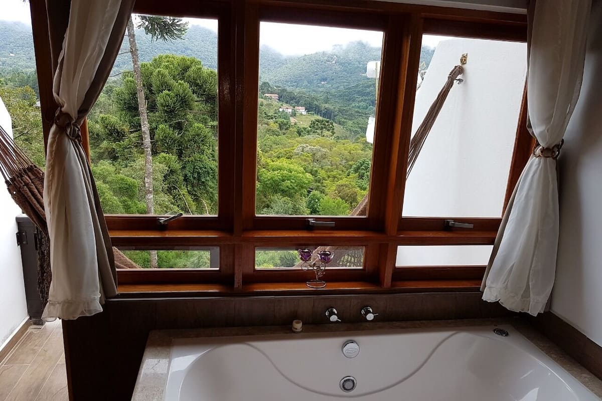 A bathroom with a large bathtub, wooden window frames, and a view of a lush green landscape.