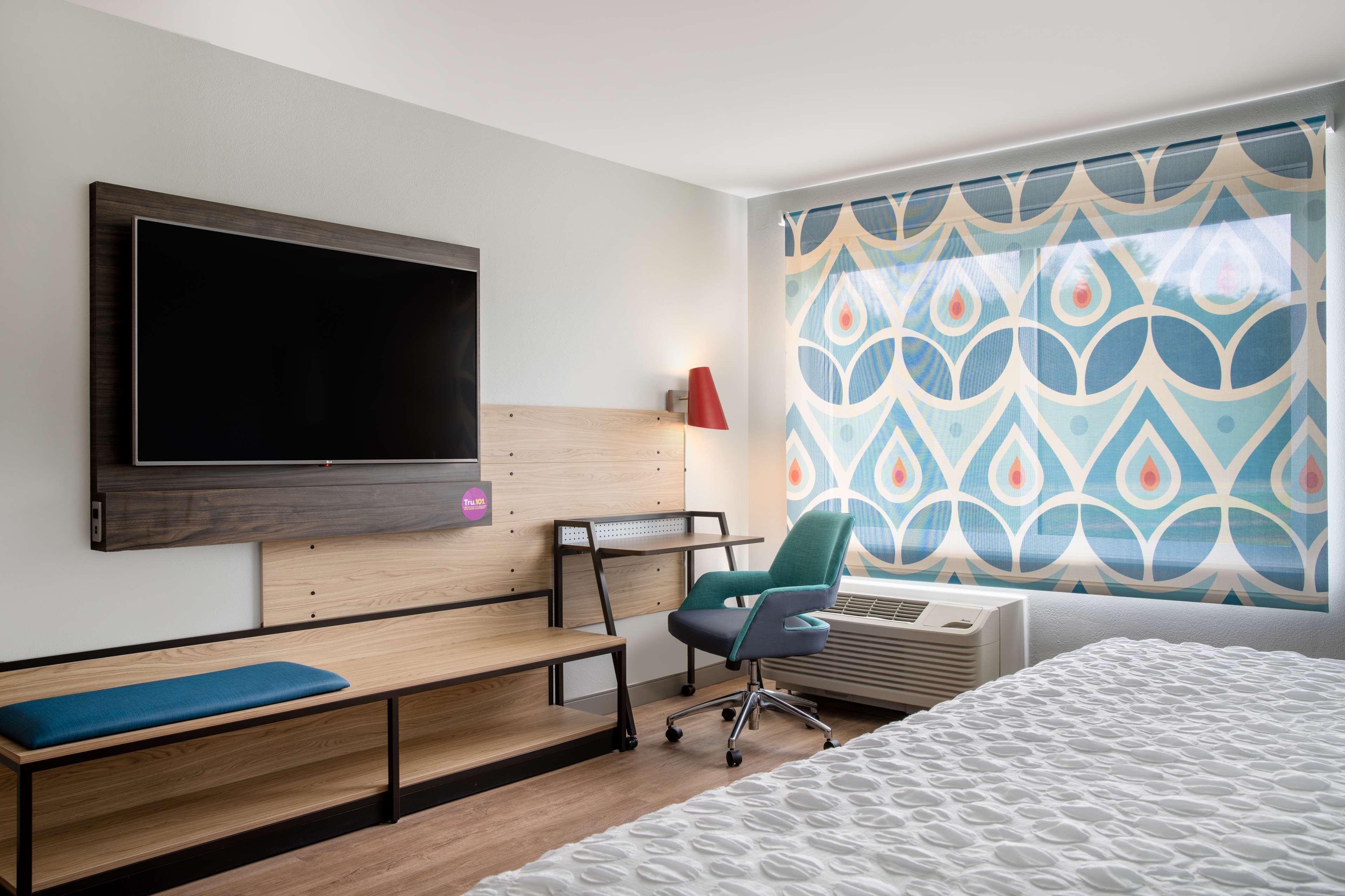 Una habitación de hotel moderna con un televisor de pantalla plana grande montado en la pared, un escritorio con silla, un banco y una cama con colcha estampada.