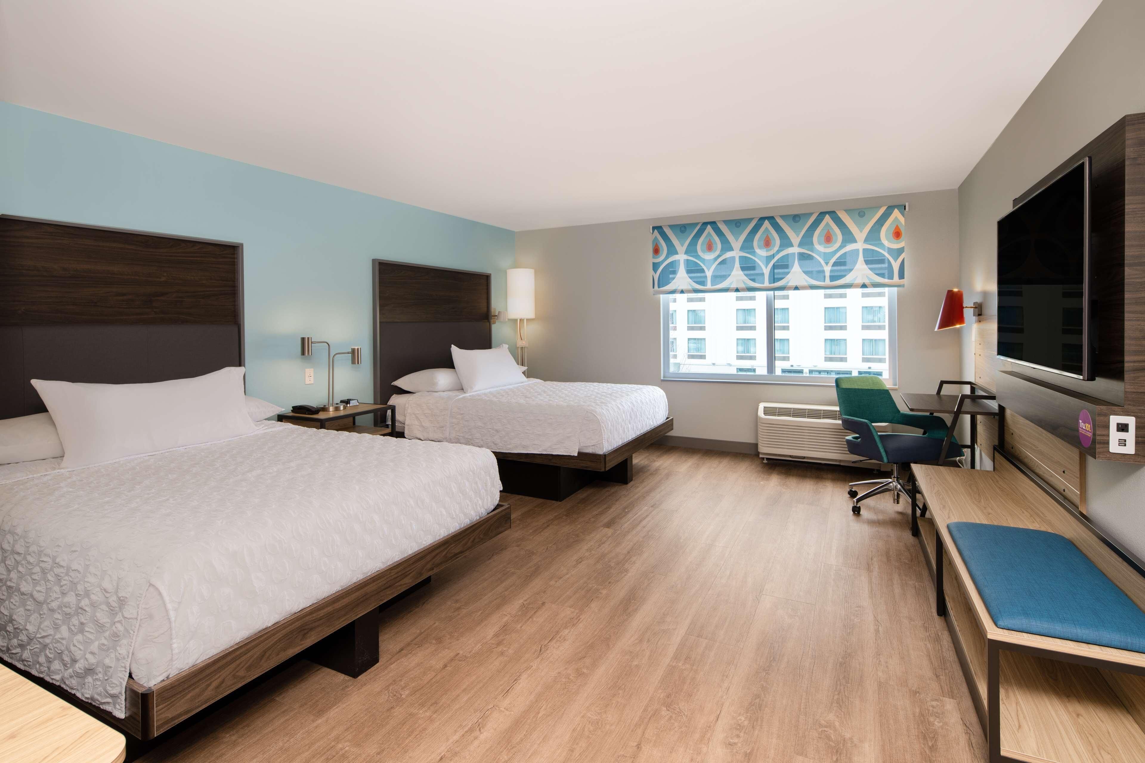 A hotel room with two beds, a desk, a chair, a TV, and a window with a view of buildings.