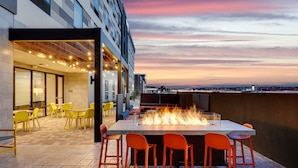 Un área de comedor al aire libre con una chimenea, sillas amarillas y vistas del horizonte de la ciudad al atardecer.