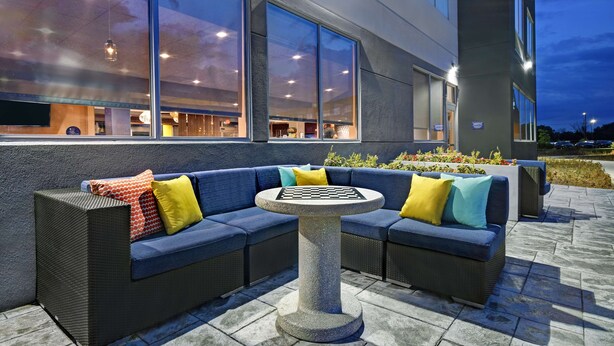 Un espace extérieur aménagé avec un canapé de couleur sombre et une table ronde, entouré de grandes fenêtres et de la façade d’un immeuble.
