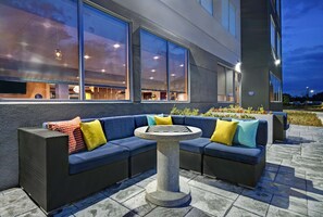 Un espace extérieur aménagé avec un canapé de couleur sombre et une table ronde, entouré de grandes fenêtres et de la façade d’un immeuble.