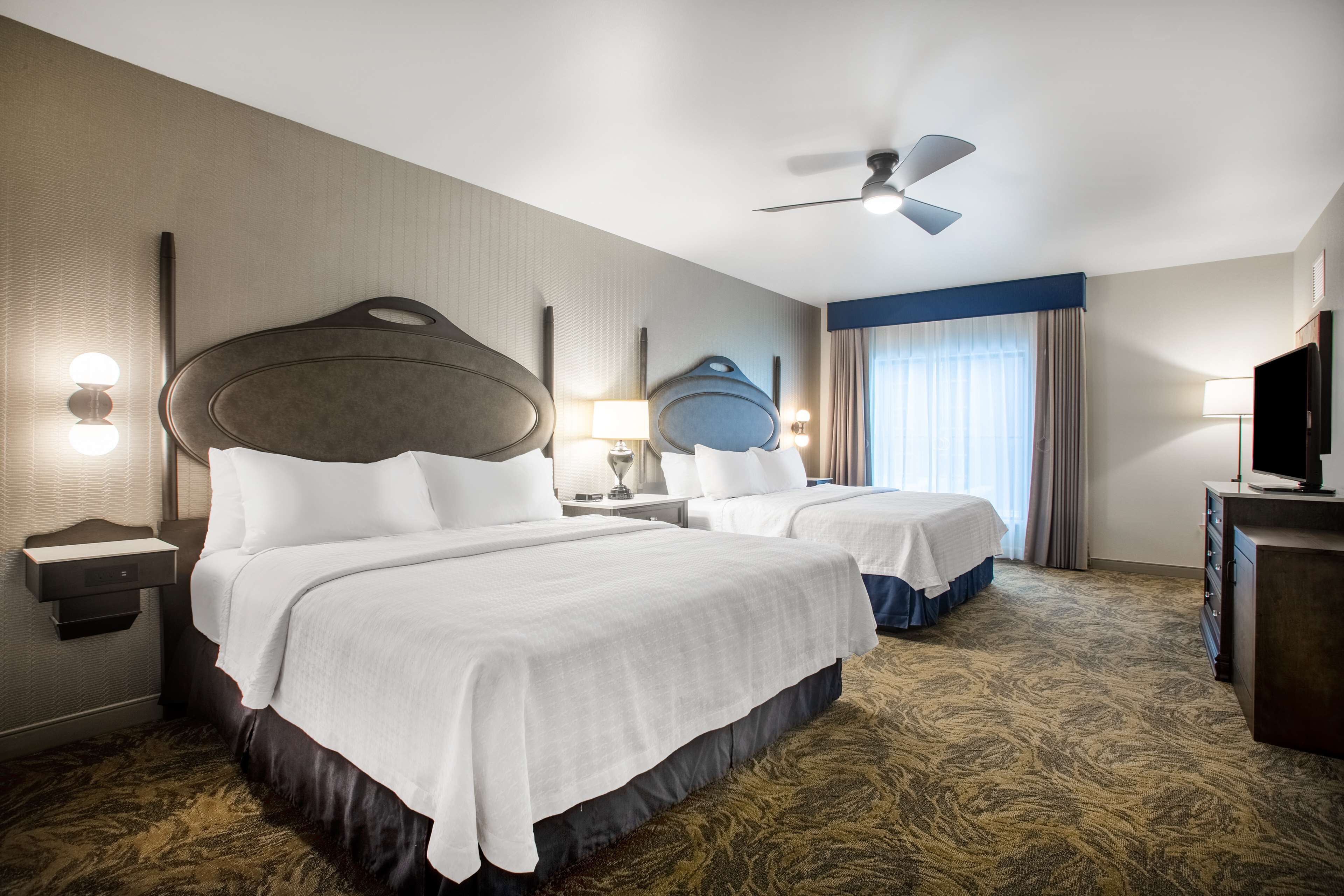 A hotel room with two beds, a ceiling fan, a TV, and a desk with a lamp.