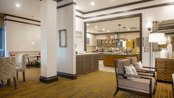 A hotel lobby with a carpeted floor, a seating area with chairs, a dining area, and a kitchen visible through an open doorway.