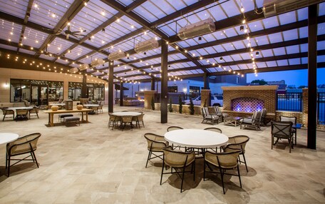An outdoor patio area with round tables and wicker chairs, a brick fireplace, and string lights.