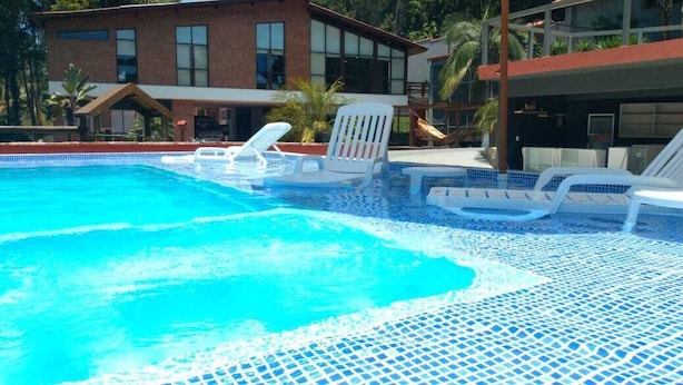 A swimming pool with clear blue water, surrounded by lounge chairs and a modern building in the background. - Pousada Vale do Sol (Embu das Artes)