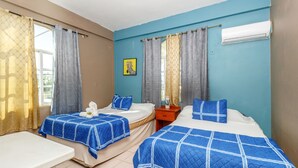 A hotel room with two beds, each with a blue quilted bedspread, a bedside table, and a nightstand. There is a window with curtains, an air conditioning unit on the wall, and a framed picture on the wall. - Mirador Hotel (Corozal)