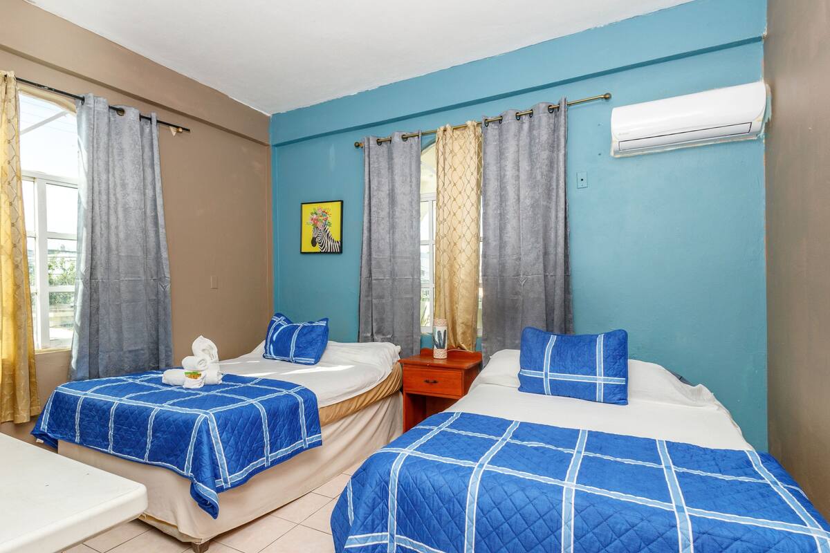 A hotel room with two beds, each with a blue quilted bedspread, a bedside table, and a nightstand. There is a window with curtains, an air conditioning unit on the wall, and a framed picture on the wall.