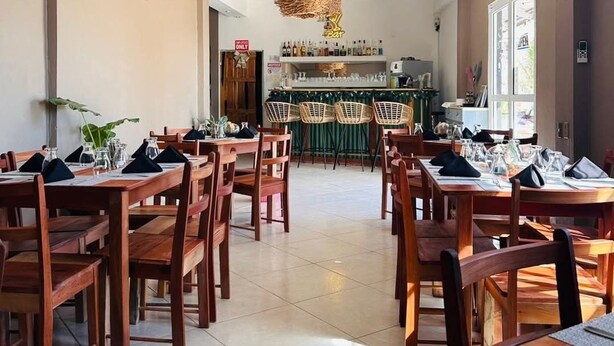 A spacious dining area with wooden tables and chairs, a central bar area with shelves of bottles, and a hanging light fixture. - Mirador Hotel (Corozal)