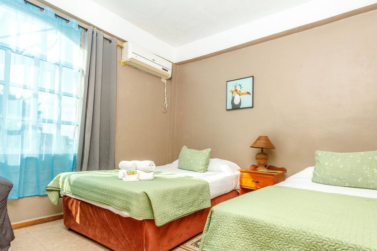 A hotel room with two beds, a window with curtains, an air conditioning unit, a bedside lamp, and a wall-mounted picture.
