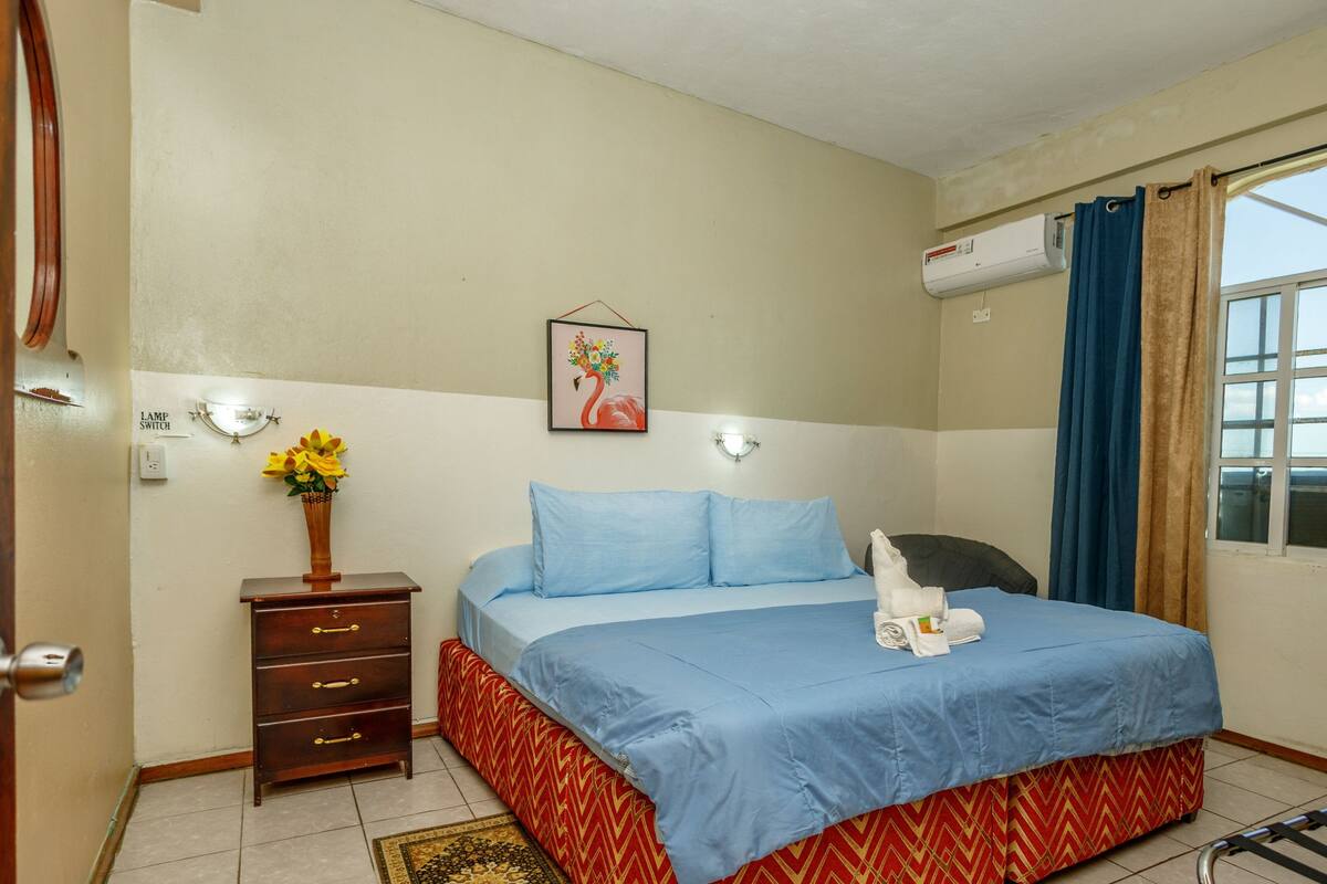 A hotel room with a bed, a dresser, a lamp switch, a wall-mounted air conditioner, and a window with curtains.