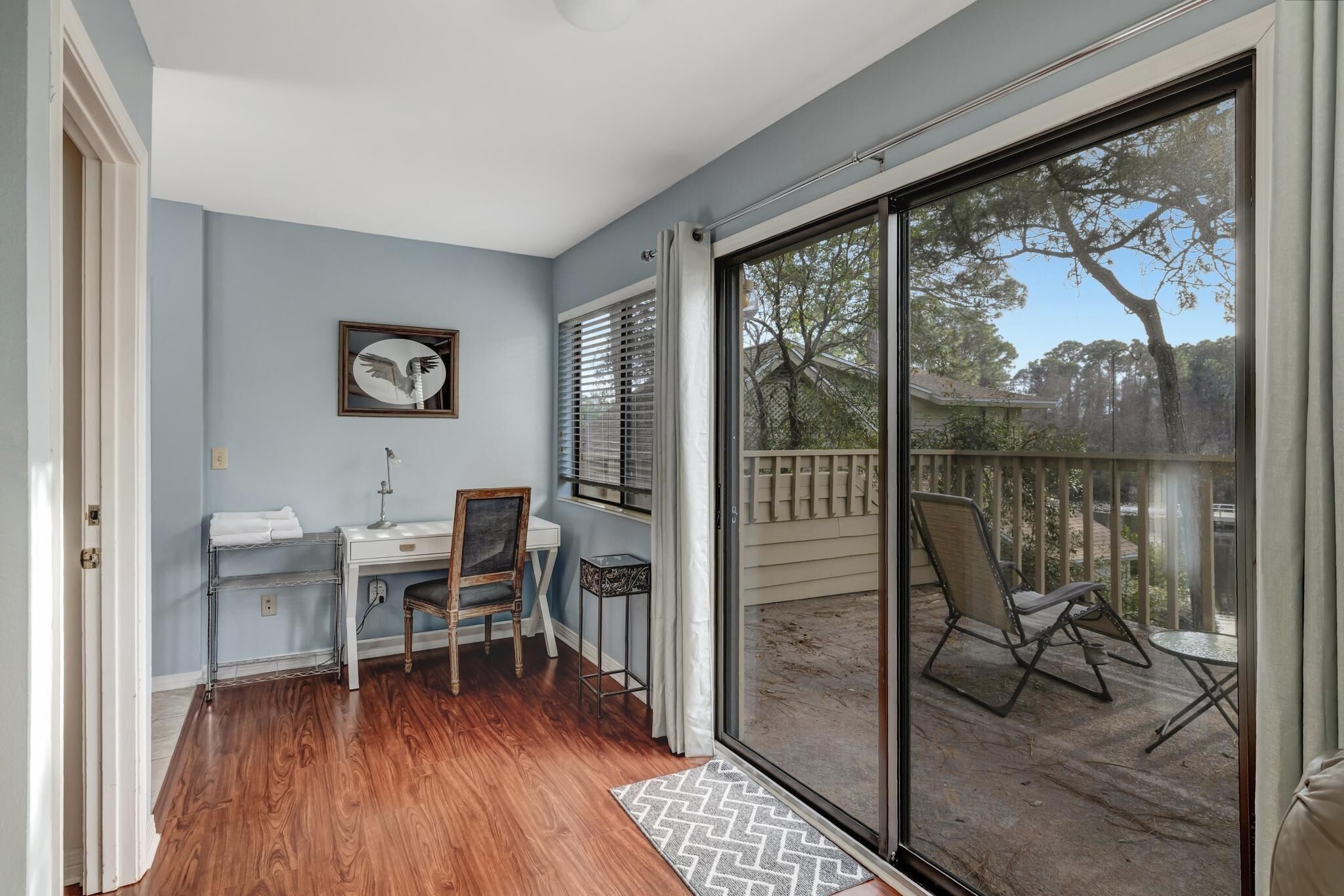 A room with a desk, chair, and a view of a balcony with outdoor furniture.