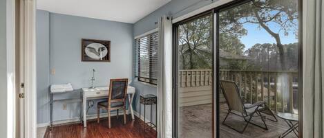 A room with a desk, chair, and a view of a balcony with outdoor furniture.