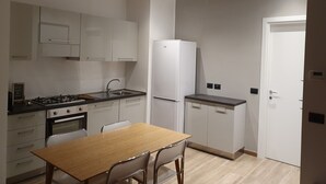 Et lille køkken med hvide skabe, indbygget ovn, køleskab og et spisebord med stole.
