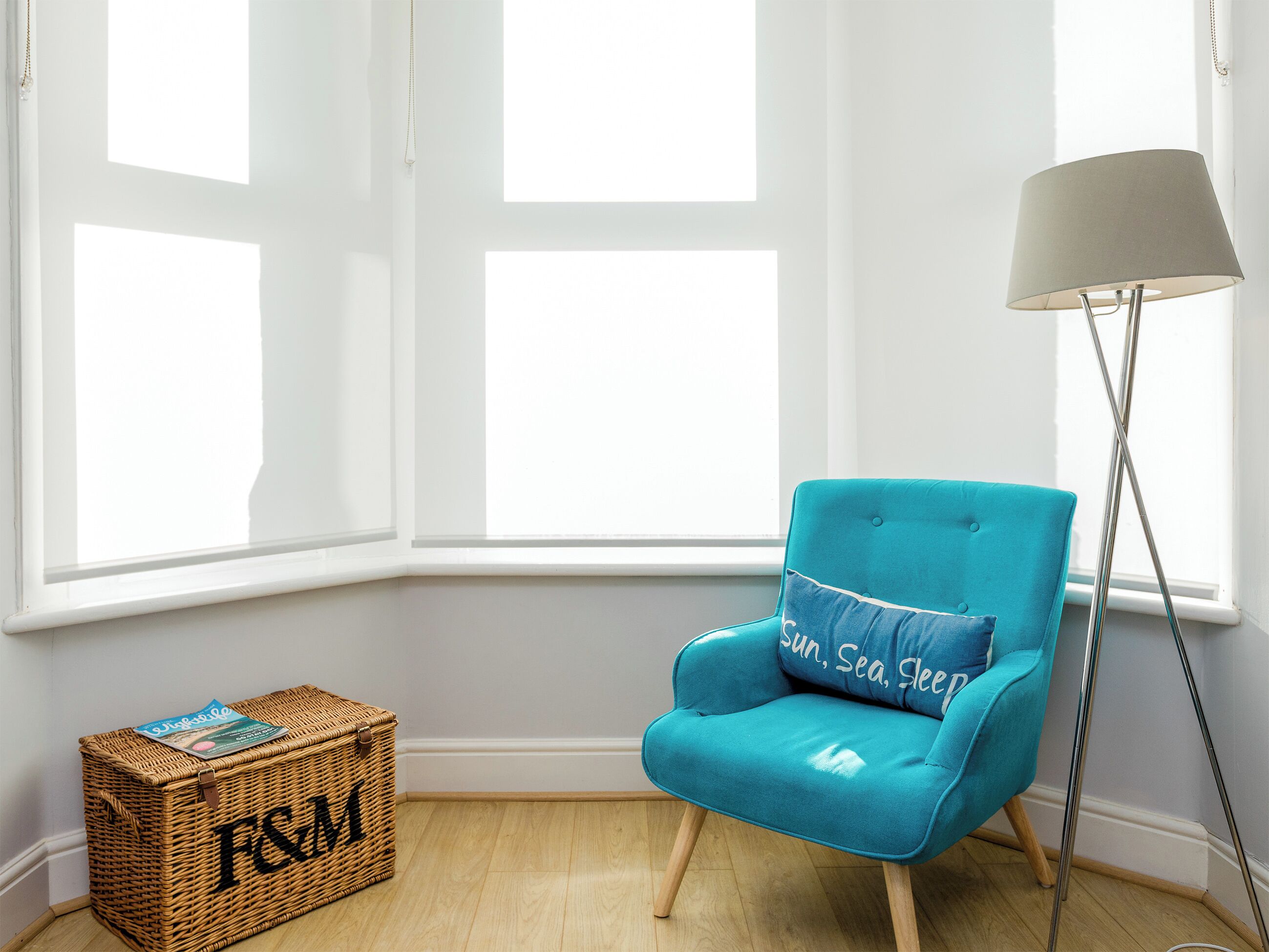 A room with a teal armchair, a floor lamp, a wicker chest of drawers, and a large window.
