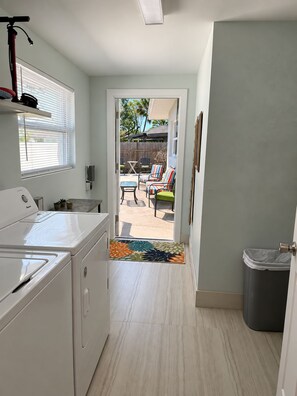 A kitchen with a washing machine, a window with blinds, and a view of an outdoor patio with a table and chairs.