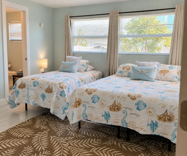 A room with two beds, each with matching bedding and pillows, a window with curtains, a lamp, and a patterned rug.