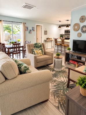 A living room with a sofa, armchairs, a coffee table, and a TV stand. There is a dining area with a table and chairs, and a kitchen in the background.