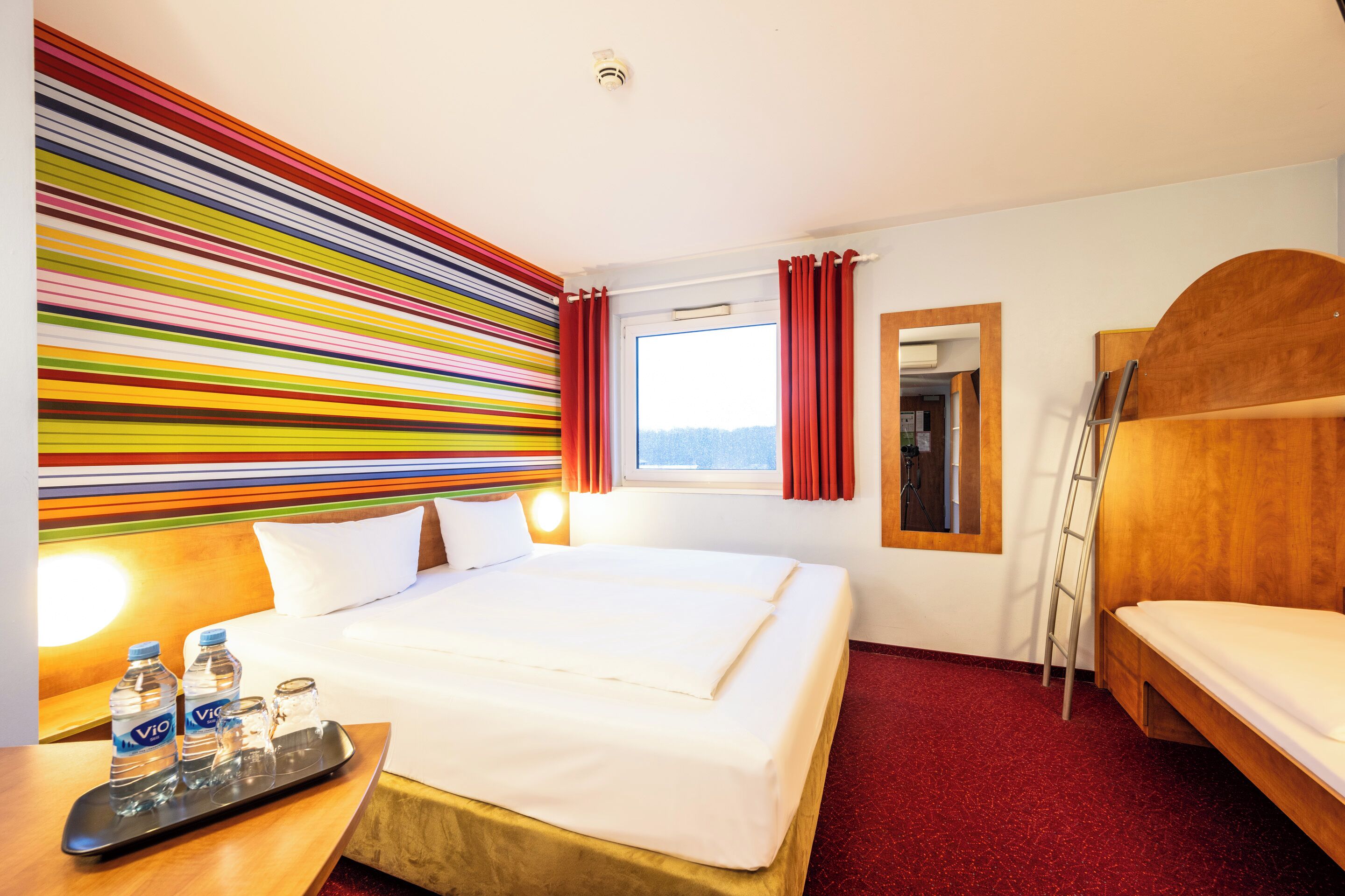 A hotel room with a large bed, a wooden bench, a small table with water bottles, and a colorful striped wall.