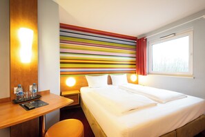 A hotel room with a bed, a desk with a chair, a nightstand, a wall with colorful stripes, and a window with curtains.