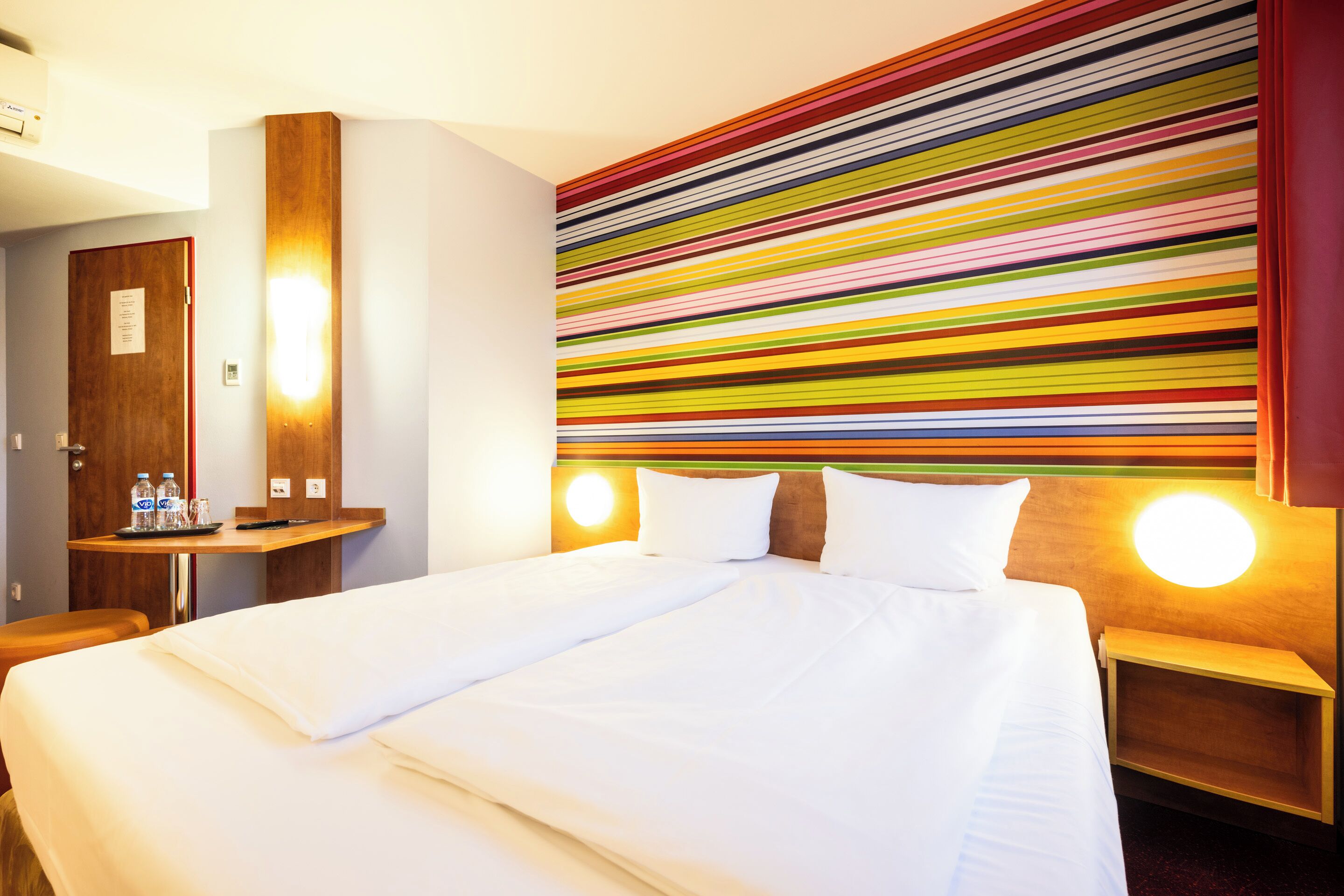 A hotel room with a large bed, a wooden nightstand, a desk with a chair, and a colorful striped wall.